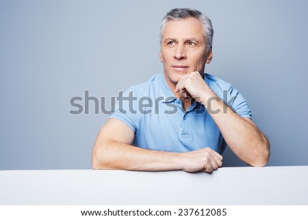 Floating away at his thoughts. Handsome senior man holding hand on chin and leaning at copy space while standing against grey background