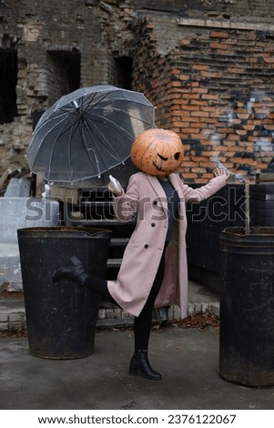 Halloween look. a girl in a light coat and with a pumpkin instead of a head stands under a transparent umbrella. There are ruins and old barrels around