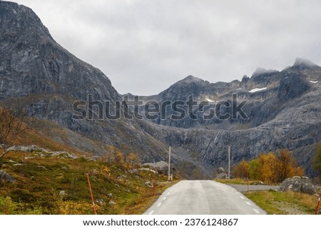 on the winding and straight road in mountains on the island of Kvaløya, in Troms, Norway. glacial valley with rocky peaks surrounded by clouds of mist in autumn. wonderful colored trees and bushes