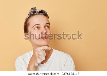 Pensive woman washes hair applies shampoo taking shower isolated over beige background looking away at advertisement area for products for hair care treatment. Royalty-Free Stock Photo #2376126113