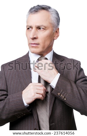 Making business look good. Confident mature man in formalwear adjusting his necktie and looking away while standing against white background