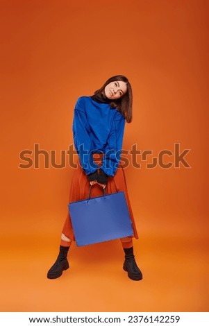 brunette woman in autumn attire standing with shopping bag on orange backdrop, black friday concept