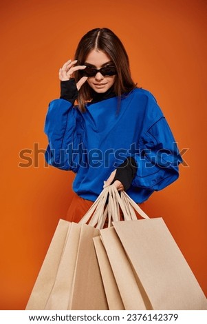 woman wearing stylish sunglasses and holding shopping bags on orange backdrop, black friday sales
