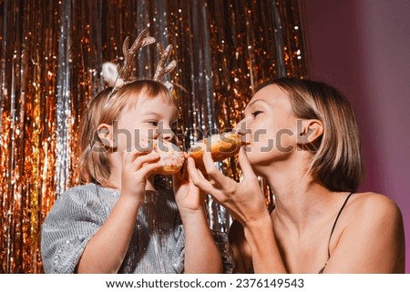 Mother and her daughter girl having fun eating donuts. Funny family with junk food on background with foil curtain decorations. Birthday party, Holiday celebration concept