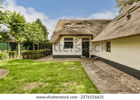 a house with thatched roof and stone pathway leading to the front door, surrounded by lush green trees on a blue sky day Royalty-Free Stock Photo #2376156631
