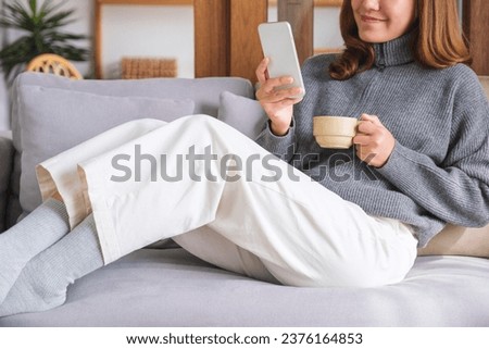 Closeup image of a young woman holding coffee cup and using smart phone while sitting on sofa at home