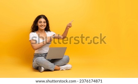 Remote job opportunities, freelance. Happy beautiful curly young indian woman student wearing casual outfit sitting on floor with laptop on her lap, pointing at copy space, yellow background, panorama Royalty-Free Stock Photo #2376174871