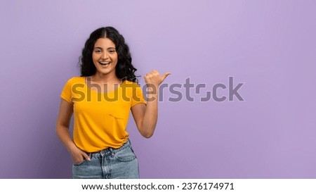 Cheerful curly young indian woman 20s wearing jeans and yellow t-shirt pointing at copy space with her thumb, showing nice offer, deal, isolated on purple background, panorama Royalty-Free Stock Photo #2376174971