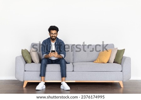 Casual Indian guy, wearing a casual outfit, sits on a cozy sofa, smiling while exploring phone, against a pristine white backdrop, illustrating a moment of joyful digital communication, copy space Royalty-Free Stock Photo #2376174995