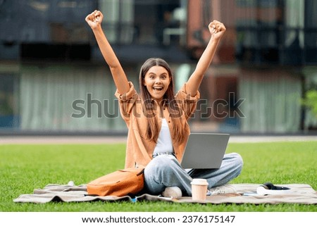 Emotional woman student with laptop on her lap raising hands up, sitting on green grass at university campus, have break between lessons, checking opportunities online, passed exam