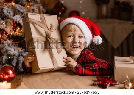 Blond boy in red plaid pajamas and a Santa hat opens a Christmas gift