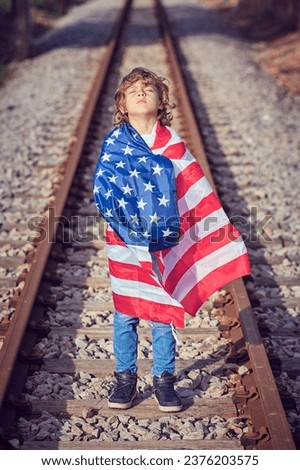 Full body of preteen boy rolled in flag of united states of america standing with closed eyes on railroad in sunny day