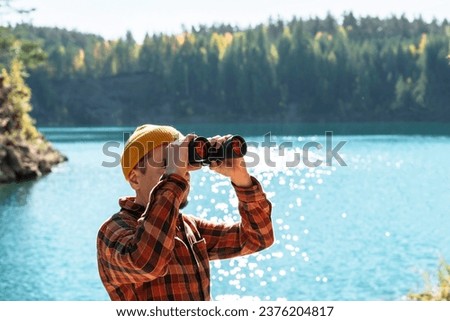 young man, dressed in red plaid shirt, engaged in birdwatching with binoculars on the banks of calm blue mountain lake in the autumn forest A haven for nature enthusiasts copy space Royalty-Free Stock Photo #2376204817