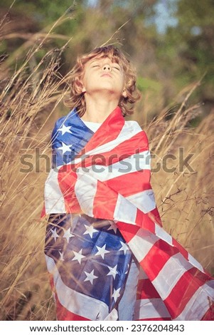 Dreamy preteen boy rolled in national stars and stripes american flag standing with closed eyes and bent head back while spending time in field