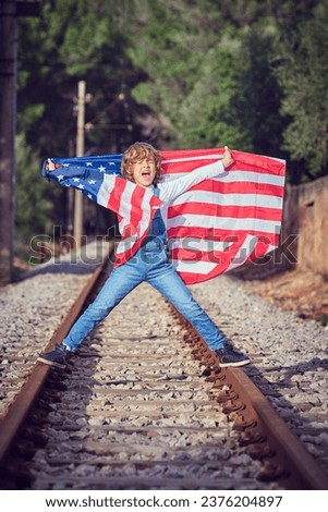 Full body of cheerful preteen boy standing on rails of railroad with flag of united states of america and screaming happily