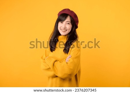 Cool young Asian woman 30s, with arms crossed on her chest, exuding a stylish vibe in a yellow sweater and red beret against a yellow background. Royalty-Free Stock Photo #2376207345