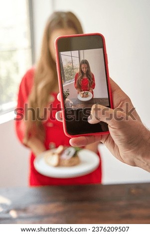 Crop anonymous person taking photo of young woman in red dress with delicious tartar while standing in kitchen
