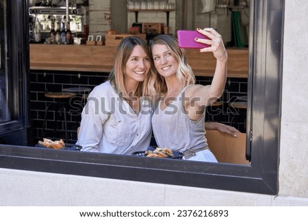 Positive young females in casual clothes taking selfie on smartphone while sitting near window and having delicious food in modern cafe