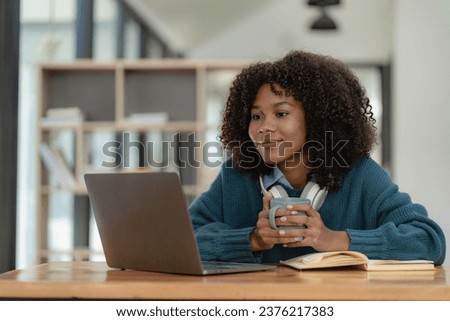 Young African American girl wearing headphones, smartphone, video call on laptop, looking at computer screen, watching webinar or doing video chat by webcam, online work concept.