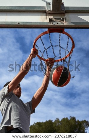 From below unrecognizable black sportsman leaping up and throwing ball into hoop while playing basketball against cloudy sky at daytime