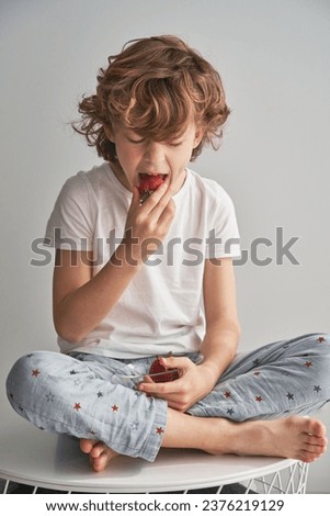 Preteen child in pajama sitting on white table and eating sweet berries at home on weekend day