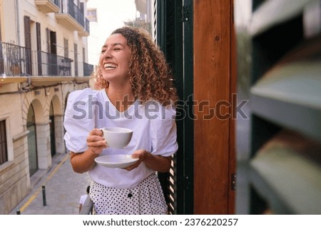 Positive young female with eyes closed laughing while drinking tea and standing on balcony over narrow city street Royalty-Free Stock Photo #2376220257