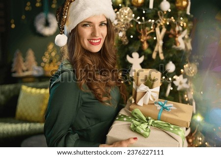 Christmas time. smiling modern woman with Santa hat and eco packaged present boxes in green dress near Christmas tree in the modern living room. Royalty-Free Stock Photo #2376227111