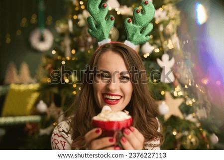 Christmas time. happy trendy housewife with festive hot chocolate cocktail and funny reindeer antlers headpiece hoop in traditional Christmas sweater near Christmas tree in the modern living room. Royalty-Free Stock Photo #2376227113