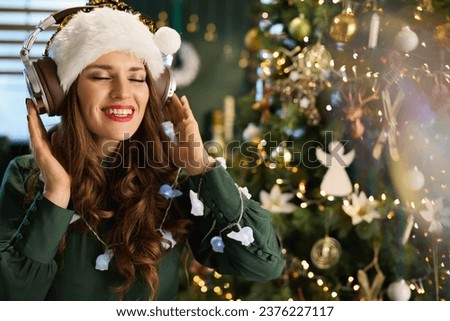Christmas time. smiling modern 40 years old housewife with Santa hat in green dress listening to the music with headphones near Christmas tree in the modern house. Royalty-Free Stock Photo #2376227117