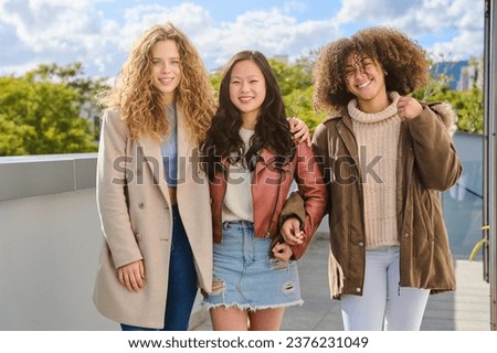 Positive young multiracial female friends in casual clothes standing on balcony in fall season and looking at camera