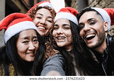 Diverse group of friends having fun celebrating Christmas on city street - Young people taking selfie picture wearing Santa Claus hats during holidays