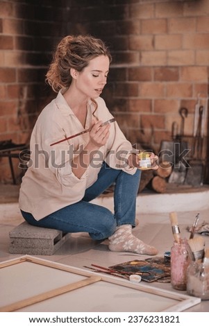 Side view of female artist sitting near fireplace and holding paintbrush and professional paint while drawing in modern studio