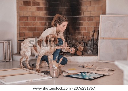 Side view of female artist with pet sitting on floor near fireplace and drawing using paintbrush and professional paints in creative workspace