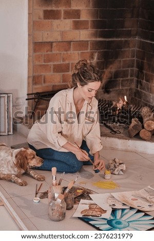 Focused female artist sitting on floor near pet and drawing on fabric using professional paintbrush and paint in creative workspace