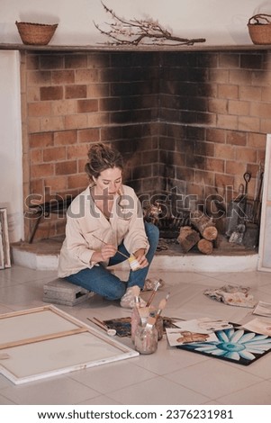 Young woman in jeans sitting on floor near fireplace with legs crossed and drawing using professional gouache paints in creative workspace