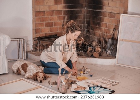 Side view of concentrated female artist in casual clothes sitting near fireplace and drawing on fabric using paintbrush and professional paint and pet sleeping near in creative workspace