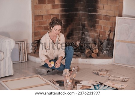 Focused female artist in casual clothes sitting on floor near fireplace and drawing using professional paints in creative workspace
