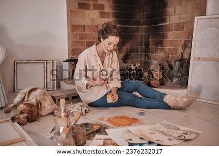 Full body of young woman in shirt and warm socks colouring paper with yellow and red gouache and looking down while spending time together with spaniel dog near burning fireplace