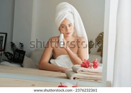 Charming woman in white towel turban sitting at table with mirror and looking at reflection while using modern vacuum pore cleaner during beauty treatment at home Royalty-Free Stock Photo #2376232361