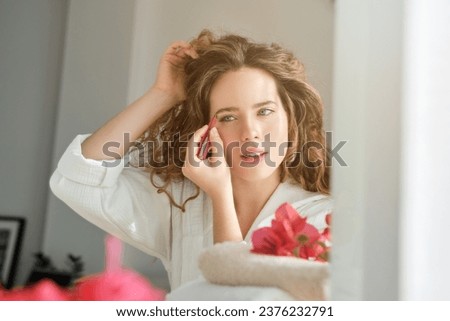 Charming female with curly hair in bathrobe plucking eyebrows while looking at reflection in mirror in light room with towel and flowers Royalty-Free Stock Photo #2376232791