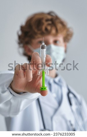 Kid in disposable mask and medical uniform with toy syringe on white background in daytime