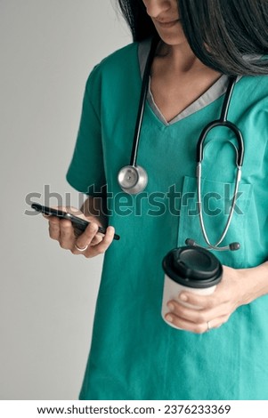 Crop unrecognizable female surgeon in green medical uniform with hot drink to go text messaging on mobile phone on light background
