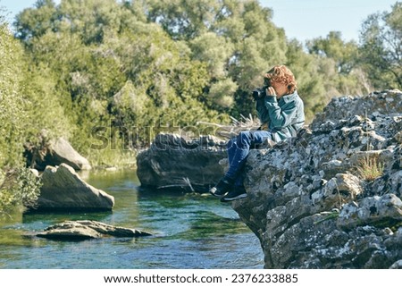Full body side view of focused boy taking picture with photo camera while sitting on edge of stony shore near flowing water in nature with green trees