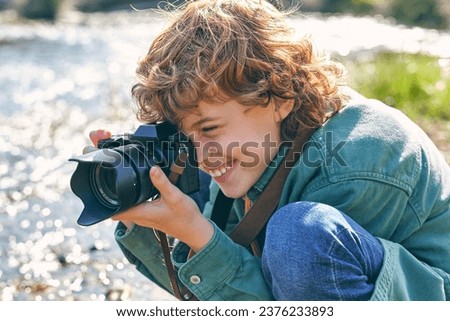 Side view of happy boy taking picture with professional photo camera while sitting on haunches near flowing river on blurred background