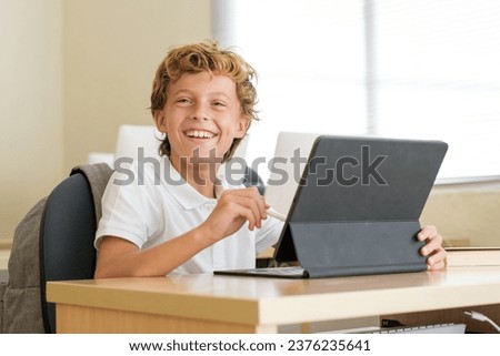 Glad schoolboy using stylus on tablet during lesson while sitting at desk in classroom at school and looking forward Royalty-Free Stock Photo #2376235641