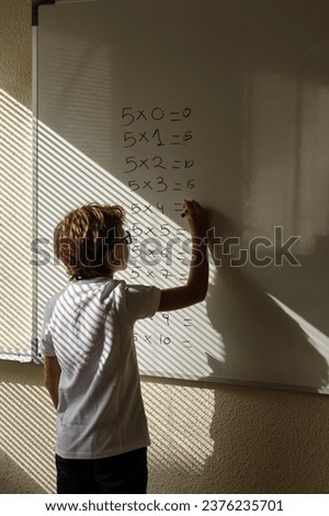 Back view of intelligent schoolboy standing writing multiplication table on whiteboard during math lesson in classroom Royalty-Free Stock Photo #2376235701