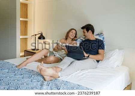 Smiling girlfriend reading book on bed near cheerful boyfriend browsing tablet while spending weekend together in bedroom Royalty-Free Stock Photo #2376239187