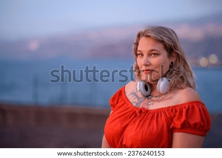 Portrait of smiling plump female in tattoo chest with red dress and headphones looking away while standing on blurred background of city Royalty-Free Stock Photo #2376240153