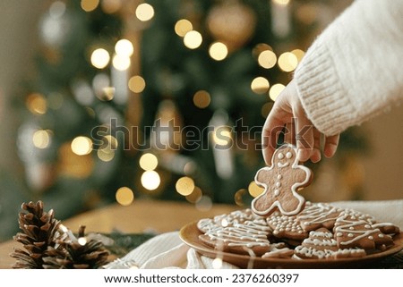 Merry Christmas! Hand holding gingerbread man cookie with icing on background of cookies in plate on table against christmas tree golden lights. Atmospheric Christmas holidays, family time