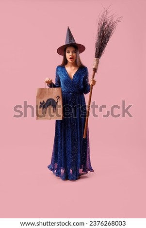 Surprised young woman dressed for Halloween as witch with broom and gift bag on pink background